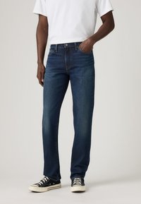 Jeans de mezclilla azul oscuro con un diseño de pierna recta, bolsillos frontales y costuras expuestas. Combinados con zapatillas de lona negras.