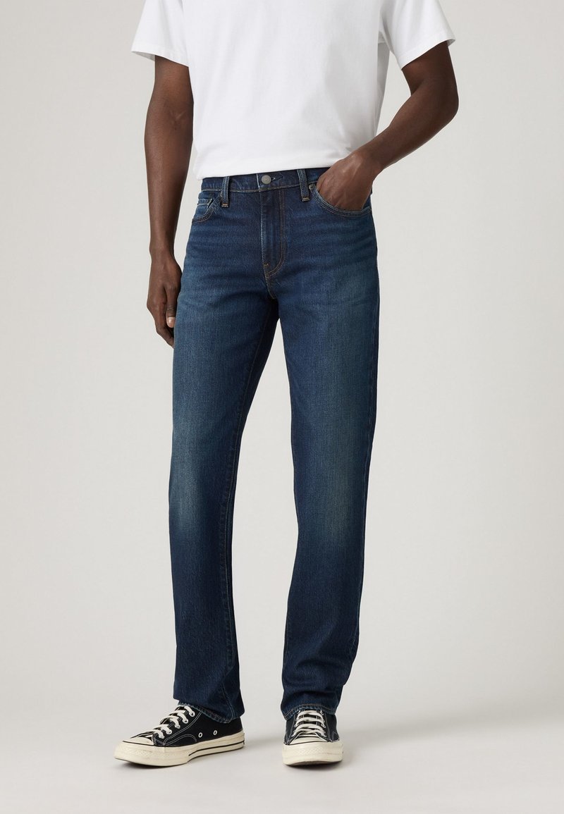 Jeans de mezclilla azul oscuro con un diseño de pierna recta, bolsillos frontales y costuras expuestas. Combinados con zapatillas de lona negras.