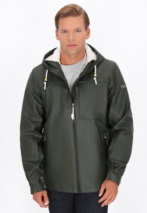 Jonge man in een donkergroene zip-up jas met een witte fleece-voering capuchon en een grijs shirt, staand tegen een witte achtergrond.