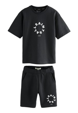 Niño de pie con las manos en los bolsillos, vistiendo camiseta y pantalones cortos negros de AllSaints, calcetines blancos de AllSaints y zapatillas blancas sobre fondo claro.