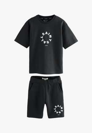 Niño de pie con las manos en los bolsillos, vistiendo camiseta y pantalones cortos negros de AllSaints, calcetines blancos de AllSaints y zapatillas blancas sobre fondo claro.