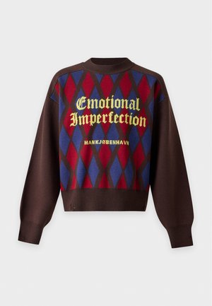 Un mannequin masculin porte une casquette grise, un pull marron à motif de losanges rouges et bleus, un pantalon noir et des chaussures marron sur un fond uni.