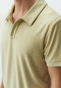 Koszulka polo z krótkim rękawem, wykonana z miękkiego, jasnobeżowego materiału. Posiada klasyczny kołnierzyk, trzy guziki oraz gładką teksturę z minimalistycznym wzornictwem.