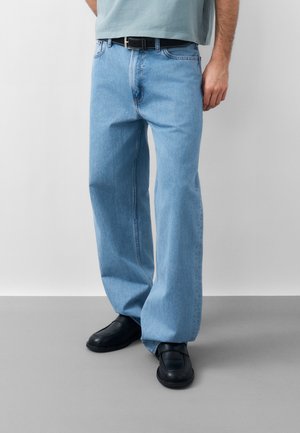 Άνδρας που φοράει ανοιχτό μπλε τζιν με φαρδιά παντελόνια, μαύρη ζώνη, μαύρα παπούτσια χωρίς κορδόνια, στέκεται μπροστά σε απλό ανοιχτό γκρι φόντο.