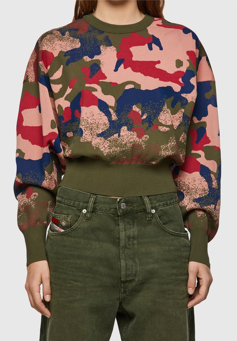 Femme portant un pull à manches longues à motif camouflage avec des nuances de rose, rouge, bleu et vert, assorti à un pantalon taille haute de couleur vert olive.