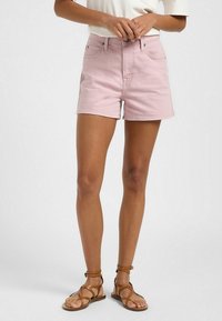 Rosa denimshorts med hög midja, femficksdesign och fransad nederkant, ihop med bruna snörsandaler, stående på en enfärgad bakgrund.