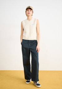 Vest crem, fără mâneci, tricotat cu fir în relief, cu decolteu în V și broderie albastră, asortat cu pantaloni largi de culoare albastră închisă și adidași multicolori.