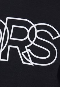 Un tejido negro presenta grandes letras blancas bordadas "ORS" con elementos de diseño interconectados, mostrando una sensación texturizada.