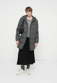 Young person wearing a gray puffer coat, gray sweater, black pleated skirt, gray socks, white sneakers, and a small black shoulder bag.