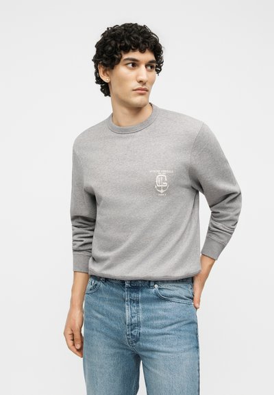 Νέος άνδρας με σγουρά μαλλιά που φοράει ανοιχτό γκρι φούτερ με λευκό λογότυπο και μεσαίου μπλε τζιν, στέκεται με το χέρι στην τσέπη.