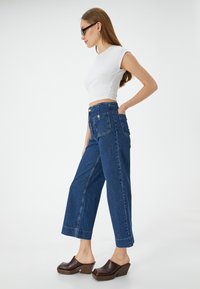 Leveitsit reilut siniset farkut, joissa on korkea vyötärö, suurilla takataskuilla ja vastakkaisella ompeleella. Yhdistetty ruskeisiin korotettuihin puukengissä.