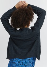 Mørkegrå strikket sweater med lange ærmer og en løs pasform, med ribbede manchetter og en høj-lav kant, båret over et blåt mønstret beklædningsstykke.