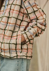 Persoană îmbrăcată într-o jachetă de fleece crem cu model în carouri multicolore și fermoar bleu deschis, cu mâna în buzunarul jachetei, asortată cu blugi albaștri.