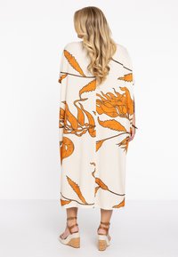 Lange jurk van crèmekleurige stof met een oranje botanisch patroon, losvallend, knielang met korte mouwen en een effen achterkant. Draagt het met sleehakken.