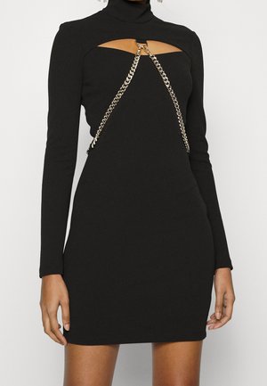 Robe mini noire à manches longues avec décolleté découpé et détail en chaîne argentée sur la poitrine, portée par une personne debout.