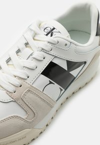 Sneaker bianca con logo e accenti neri, caratterizzata da un pannello in suede, suola testurizzata e lacci bianchi. Materiali lisci e a contrasto.