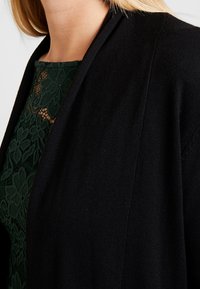 Svart kofta med öppen fram, gjord av ett mjukt stickat tyg. Under finns en mörkgrön spetsblus med blommönster och genomskinlig textur.