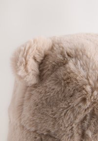 Apvalus, smėlio spalvos minkštas audinys su švelnia, pūkuota tekstūra. Viršuje yra mažos, apvalios ausytės, suteikiančios jaukų ir viliojantį vaizdą.