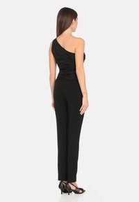 Top nero a spalla unica con texture arricciata, abbinato a pantaloni neri aderenti. La modella indossa tacchi neri a fascia. Sfondo bianco semplice.