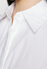 Camicia bianca con colletto classico, tessuto liscio e due bottoni visibili allo scollo.