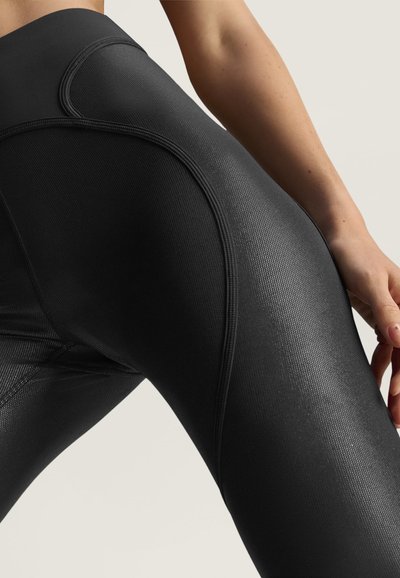 Close-up af en person iført teksturerede sorte leggings med buede sømdetaljer og en hånd hvilende nær låret.