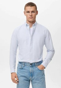 Camisa de botones a rayas azul claro y blanca con mangas largas, llevada sobre una camiseta blanca, combinada con jeans de mezclilla azul claro.