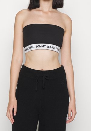 Kvinde iført en sort tubetop med et hvidt bånd med teksten "Tommy Jeans" og sorte bukser med snøre på en ensfarvet baggrund.