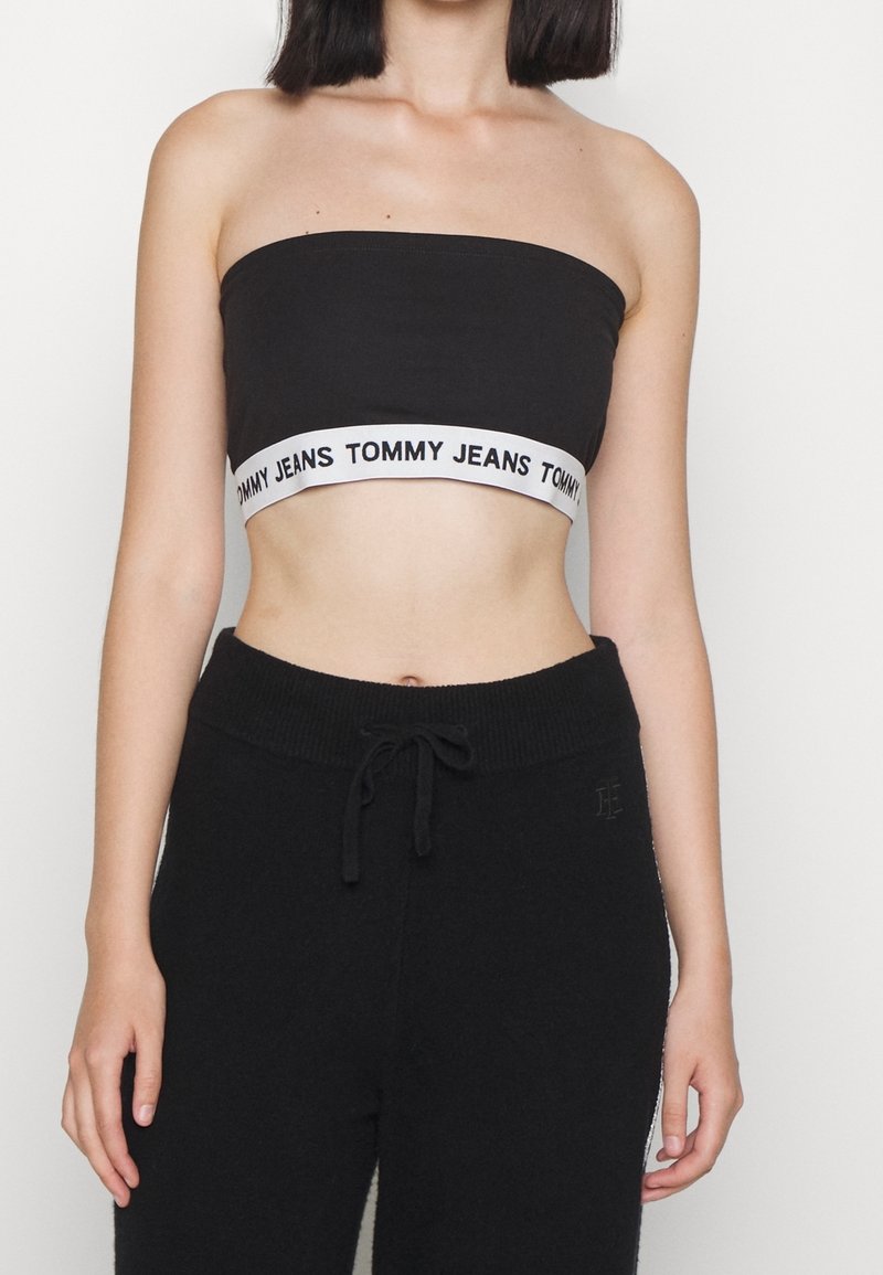 Femme portant un haut bandeau noir avec une bande blanche portant l'inscription "Tommy Jeans" et un pantalon noir à cordons sur un fond uni.