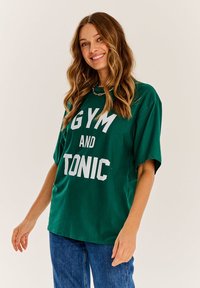 Zelené nadrozmerné tričko vyrobené z bavlny. S jasným bielym nápisom "GYM AND TONIC." Krátke rukávy a voľný strih.