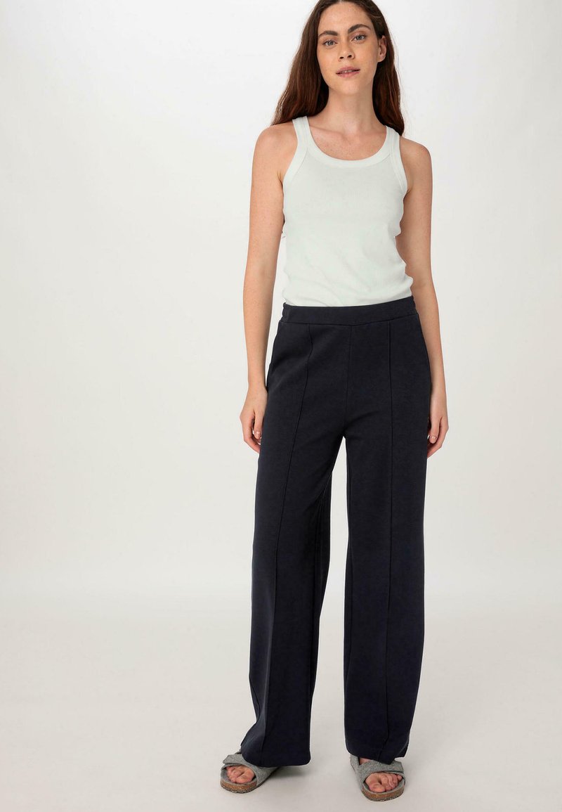 Donkerblauwe wijde broek met een gladde textuur en voornaden, gecombineerd met een lichtgrijze tanktop. Schoeisel is een paar grijze instapsandalen.