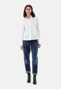 Camicia bianca con maniche lunghe, abbinata a jeans blu scuro con risvolti e stivaletti neri a caviglia, su uno sfondo grigio chiaro.