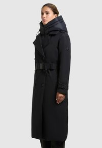 Zwarte trenchcoat met een afneembare gewatteerde capuchon, een riem in de taille, dubbel geknoopte knopen en een gestructureerde stof. Voorzien van zijzakken.