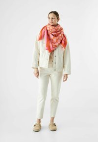 Großer rosafarbener Schal mit abstraktem Herzmuster, über eine cremefarbene Jeansjacke drapiert, kombiniert mit cremefarbenen Hosen und beigen Slippern.