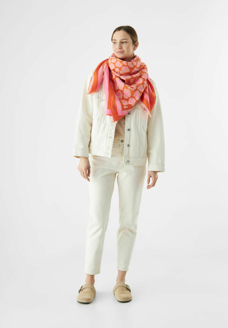 Großer rosafarbener Schal mit abstraktem Herzmuster, über eine cremefarbene Jeansjacke drapiert, kombiniert mit cremefarbenen Hosen und beigen Slippern.