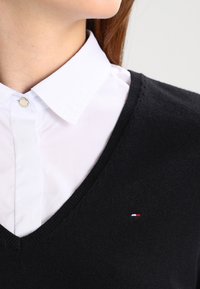 Schwarzer V-Ausschnitt-Pullover über einem weißen Hemd mit Kragen, mit einem kleinen rot-weiß-blauen Logo auf der linken Seite. Glatte Textur.