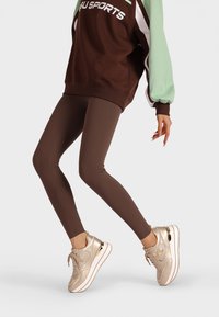 Leggings atletici marroni abbinati a una felpa oversize marrone e menta. Le calzature presentano dettagli dorati su sneakers crema con un design in rete.