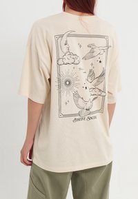 Camiseta de gran tamaño en tono beige con un diseño gráfico negro de nubes, una luna, pájaros, estrellas y el texto "Positive Sincere" en la parte posterior.