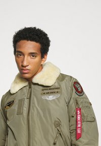 Olivgrüne Bomberjacke mit einem Kunstfellkragen, bestickten Aufnähern und einem roten "Remove Before Flight"-Tag. Mit Reißverschlusstaschen; glatte Textur.