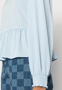 Light blue blouse with gathered waistline and elasticized cuffs. Paired with blue checkered patterned pants in varying shades of blue.