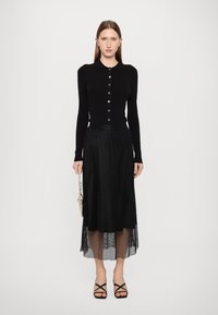 Cardigã preto de malha com gola e fecho de botão, combinado com uma saia preta de malha transparente. A modelo segura uma pequena bolsa bege.