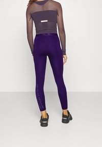 Leggings morados con una textura suave, cinturilla alta y texto gráfico blanco a lo largo del costado, combinados con una camiseta de malla de manga larga oscura.