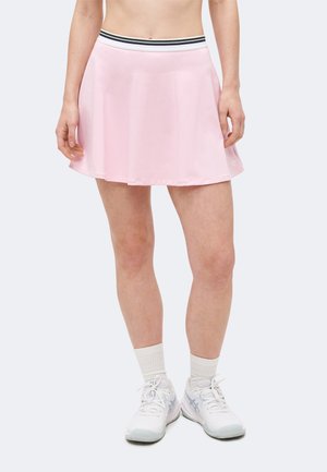 Γυναίκα που φοράει αθλητική φούστα ανοιχτό ροζ με μαύρη και λευκή μέση, λευκές κάλτσες και λευκά αθλητικά παπούτσια, στέκεται μπροστά από ανοιχτό φόντο.