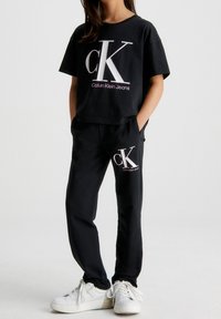 Černé zkrácené tričko s velkým bílým logem "ck" a růžovým textem, spárované s černými teplákovými kalhotami se stejným logem, nošené s bílými teniskami.