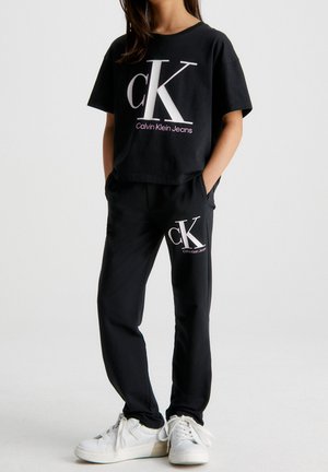 Czarny krótki T-shirt z dużym białym logo "ck" oraz różowym napisem, w połączeniu z czarnymi spodniami dresowymi z pasującym logo, noszony z białymi sneakersami.