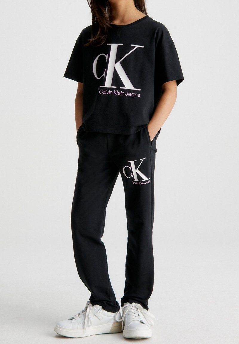 Černé zkrácené tričko s velkým bílým logem "ck" a růžovým textem, spárované s černými teplákovými kalhotami se stejným logem, nošené s bílými teniskami.