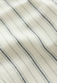 Tissu avec un fond crème présentant de fines rayures verticales noires. Le tissu a un tissage texturé avec des variations de motif subtiles.