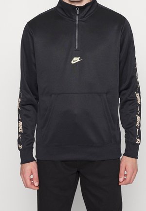 Osoba ubrana w czarną bluzę Nike z zamkiem do połowy długości i kieszenią z przodu oraz beżowymi logo Nike na obu rękawach.