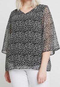 Femme portant un chemisier noir transparent avec des points blancs irréguliers et des manches larges, associé à un pantalon blanc, sur un fond uni.