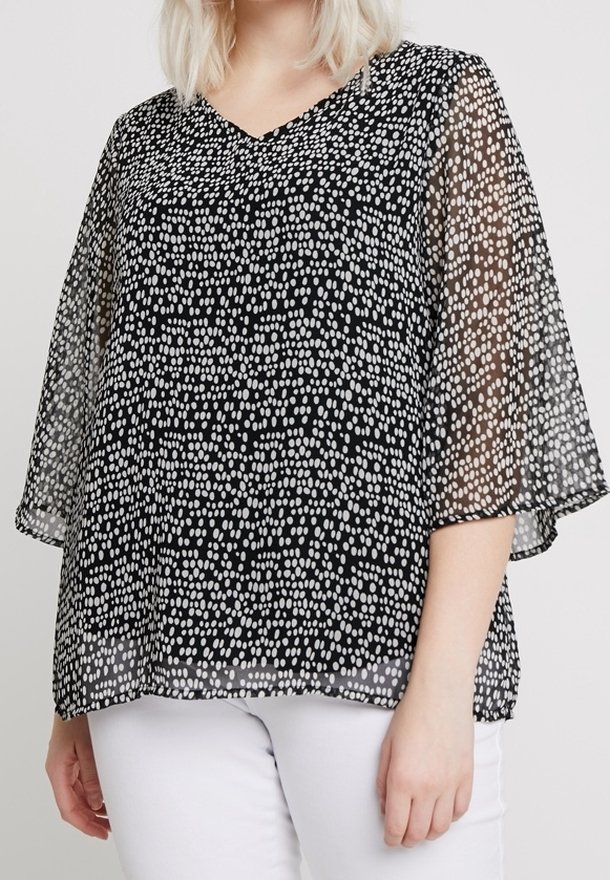 Femme portant un chemisier noir transparent avec des points blancs irréguliers et des manches larges, associé à un pantalon blanc, sur un fond uni.