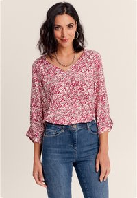 Rot-weiße geblümte Bluse mit V-Ausschnitt, Dreiviertelärmeln und gerafften Details an der Vorderseite, kombiniert mit blauen Jeans.